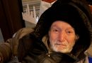 Homme âgé emmitouflé dans un manteau épais, assis à l’intérieur d’un abri urbain, le visage marqué par la fatigue et le froid, regard humide et épuisé.