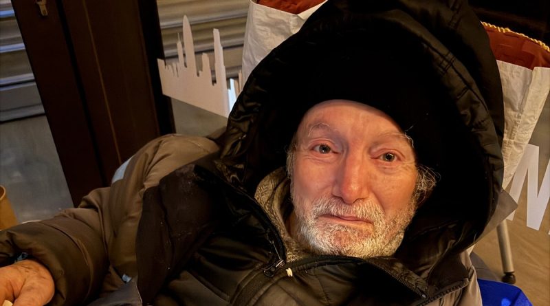 Homme âgé emmitouflé dans un manteau épais, assis à l’intérieur d’un abri urbain, le visage marqué par la fatigue et le froid, regard humide et épuisé.