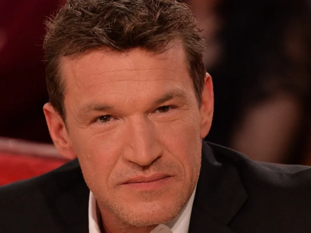 Homme d’âge mûr aux cheveux courts bruns, regard sérieux, assis sur un plateau de télévision avec un fond flou et des lumières chaudes