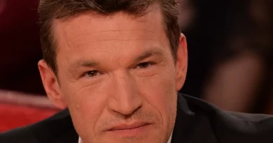 Homme d’âge mûr aux cheveux courts bruns, regard sérieux, assis sur un plateau de télévision avec un fond flou et des lumières chaudes