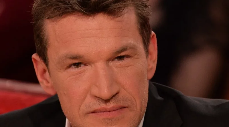 Homme d’âge mûr aux cheveux courts bruns, regard sérieux, assis sur un plateau de télévision avec un fond flou et des lumières chaudes