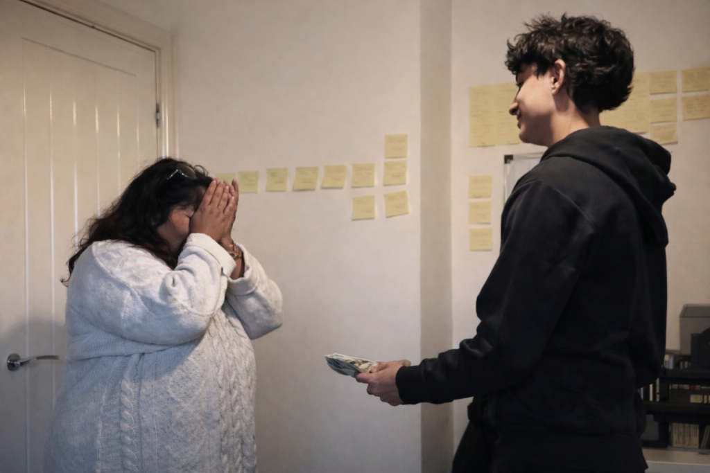 Un adolescent tend une liasse de billets à sa mère bouleversée, qui se couvre le visage d’émotion, dans une pièce simple et lumineuse.