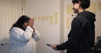 Un adolescent tend une liasse de billets à sa mère bouleversée, qui se couvre le visage d’émotion, dans une pièce simple et lumineuse.