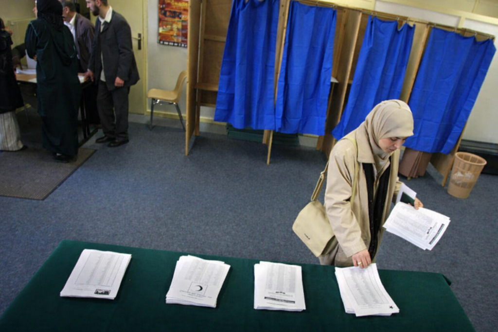 Femme portant un voile sélectionnant un bulletin de vote dans un bureau de vote municipal, avec des piles de bulletins sur une table et des isoloirs bleus en arrière-plan