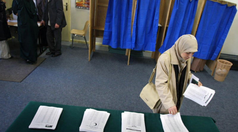 Femme portant un voile sélectionnant un bulletin de vote dans un bureau de vote municipal, avec des piles de bulletins sur une table et des isoloirs bleus en arrière-plan