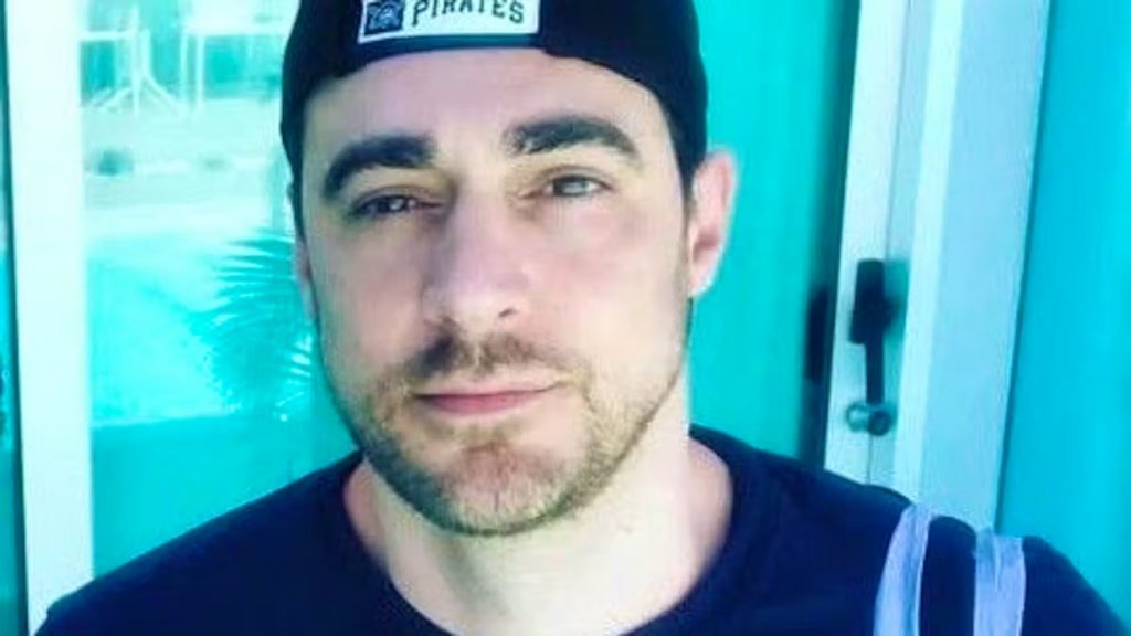 Portrait d’un homme portant une casquette sombre, regard posé vers l’objectif, photographié en gros plan devant une baie vitrée laissant apparaître un décor extérieur lumineux.
