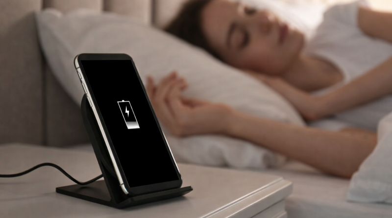 Smartphone en charge posé sur une table de nuit à côté d’un lit dans une chambre sombre.