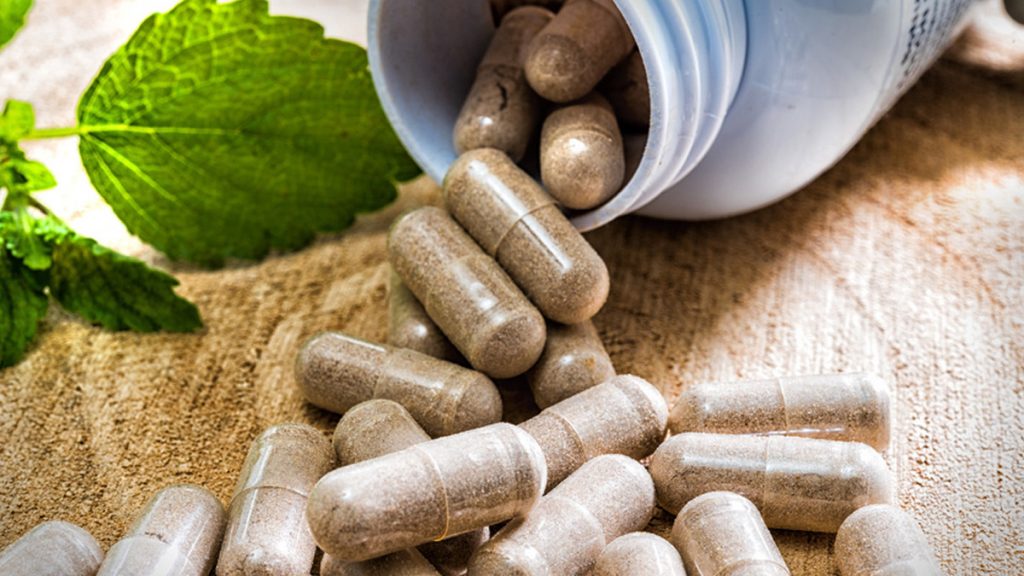gélules de compléments alimentaires renversées hors d’un flacon, posées sur une surface naturelle avec des feuilles vertes en arrière-plan.