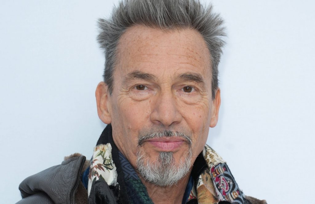 Portrait en gros plan d’un chanteur français aux cheveux gris dressés, barbe poivre et sel, regard sérieux face à l’objectif, sur fond clair.