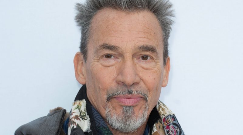 Portrait en gros plan d’un chanteur français aux cheveux gris dressés, barbe poivre et sel, regard sérieux face à l’objectif, sur fond clair.