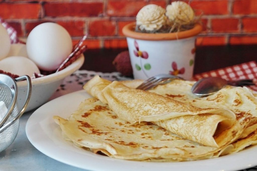 Crêpes fines et dorées empilées dans une assiette blanche, posées sur une table avec des œufs et des ustensiles de cuisine, dans une ambiance chaleureuse évoquant une tradition culinaire hivernale.