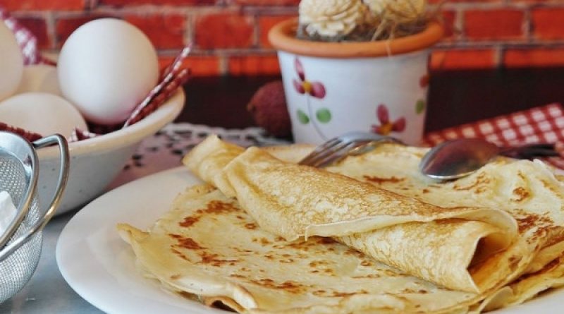 Crêpes fines et dorées empilées dans une assiette blanche, posées sur une table avec des œufs et des ustensiles de cuisine, dans une ambiance chaleureuse évoquant une tradition culinaire hivernale.