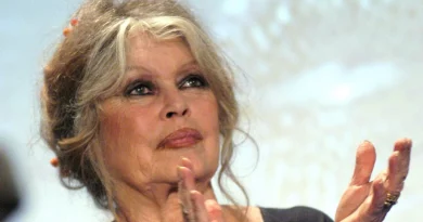 Femme âgée aux cheveux clairs coiffés en chignon lâche, maquillage prononcé, applaudissant lors d’un événement public dans une ambiance solennelle.