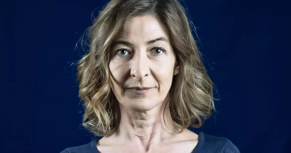 Portrait serré d’une femme aux cheveux châtains ondulés, regard sérieux, sur fond bleu sombre, éclairage studio mettant en valeur les traits du visage.