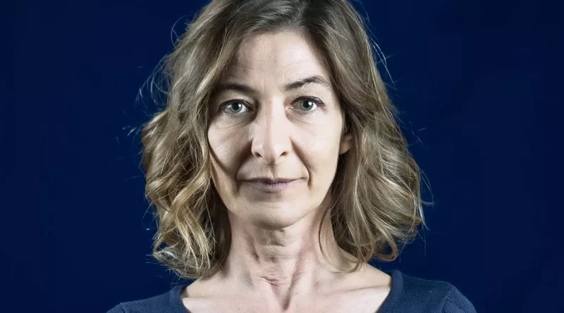 Portrait serré d’une femme aux cheveux châtains ondulés, regard sérieux, sur fond bleu sombre, éclairage studio mettant en valeur les traits du visage.