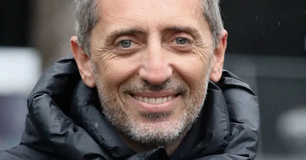 Portrait en gros plan d’un homme d’âge mûr souriant, portant une veste sombre rembourrée, avec un regard direct et une expression sereine, en extérieur.