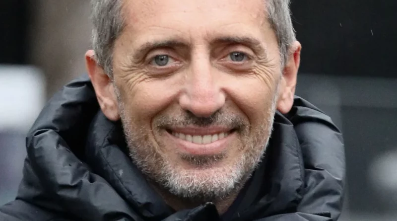 Portrait en gros plan d’un homme d’âge mûr souriant, portant une veste sombre rembourrée, avec un regard direct et une expression sereine, en extérieur.