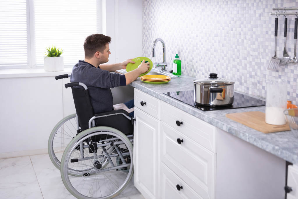 Un homme en fauteuil roulant lave de la vaisselle dans une cuisine adaptée, illustrant une situation de vie quotidienne autonome dans un logement moderne.