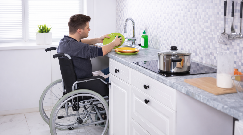 Un homme en fauteuil roulant lave de la vaisselle dans une cuisine adaptée, illustrant une situation de vie quotidienne autonome dans un logement moderne.