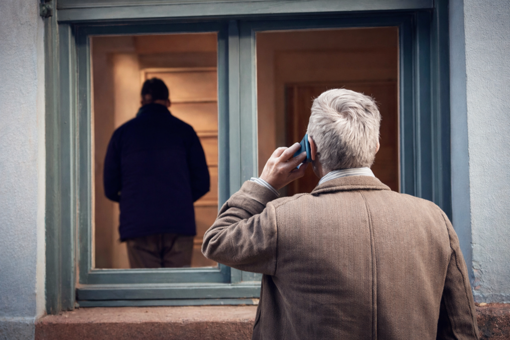 Un propriétaire âgé téléphone devant la fenêtre de son logement occupé illégalement, observant un homme à l’intérieur d’un appartement vide, illustrant la détresse face au squat.