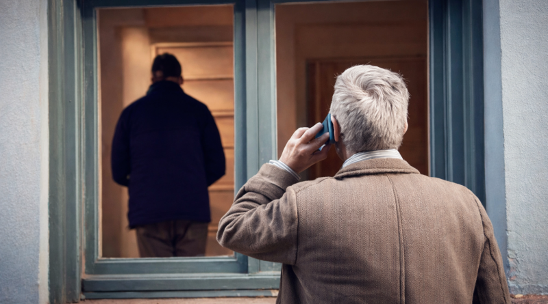 Un propriétaire âgé téléphone devant la fenêtre de son logement occupé illégalement, observant un homme à l’intérieur d’un appartement vide, illustrant la détresse face au squat.