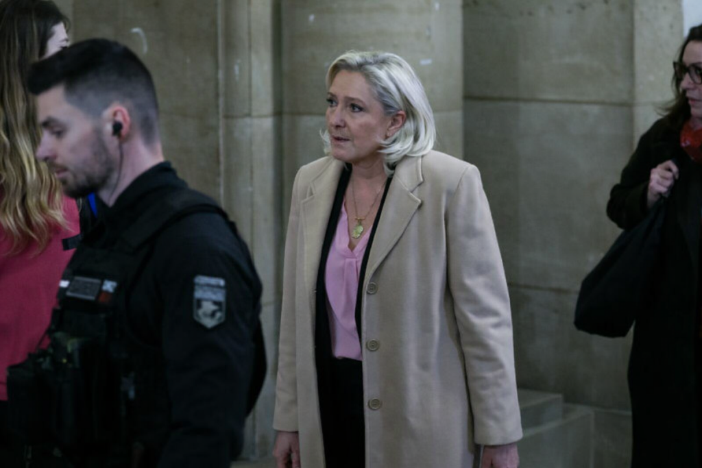 Femme blonde marchant dans un couloir de tribunal ancien, entourée d’agents et de collaborateurs, ambiance solennelle et judiciaire.