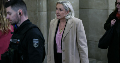 Femme blonde marchant dans un couloir de tribunal ancien, entourée d’agents et de collaborateurs, ambiance solennelle et judiciaire.
