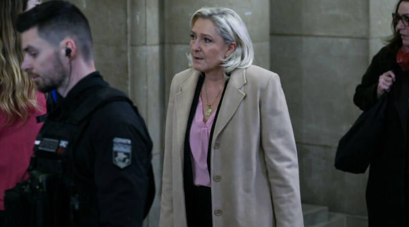 Femme blonde marchant dans un couloir de tribunal ancien, entourée d’agents et de collaborateurs, ambiance solennelle et judiciaire.