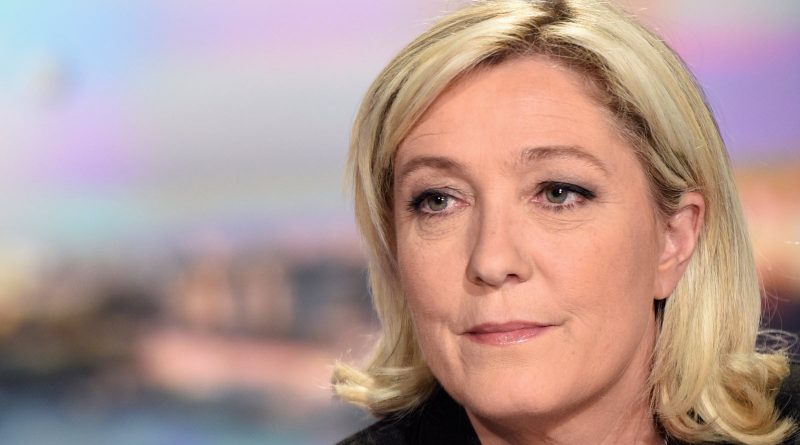 Portrait serré d’une femme politique blonde, expression grave, regard fixe, fond flou aux tons pastel évoquant un plateau télévisé