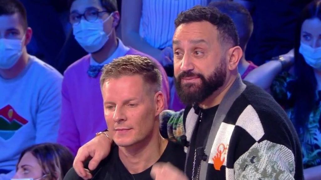 deux animateurs de télévision assis côte à côte sur un plateau, l’un posant une main sur l’épaule de l’autre, sous les projecteurs, avec un public en arrière-plan