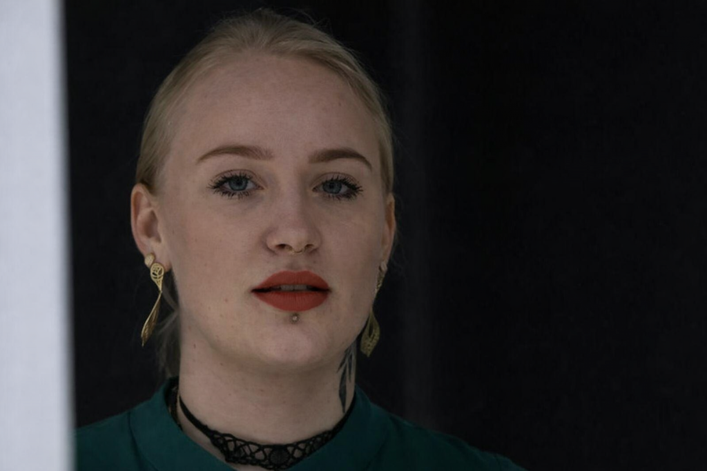 Portrait serré d’une jeune femme blonde regardant droit devant elle, lèvres rouges, bijoux discrets et piercing, sur fond sombre flouté, ambiance sérieuse et journalistique.