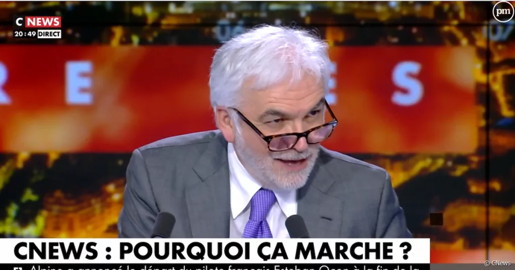 Animateur de télévision aux cheveux blancs et à la barbe grise, portant un costume et une cravate, s’exprimant en plateau lors d’une émission de débat en direct