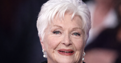 Portrait rapproché d’une femme âgée aux cheveux blancs, maquillage discret et sourire apaisant, photographiée lors d’un événement public avec un arrière-plan flouté