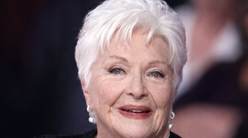 Portrait rapproché d’une femme âgée aux cheveux blancs, maquillage discret et sourire apaisant, photographiée lors d’un événement public avec un arrière-plan flouté