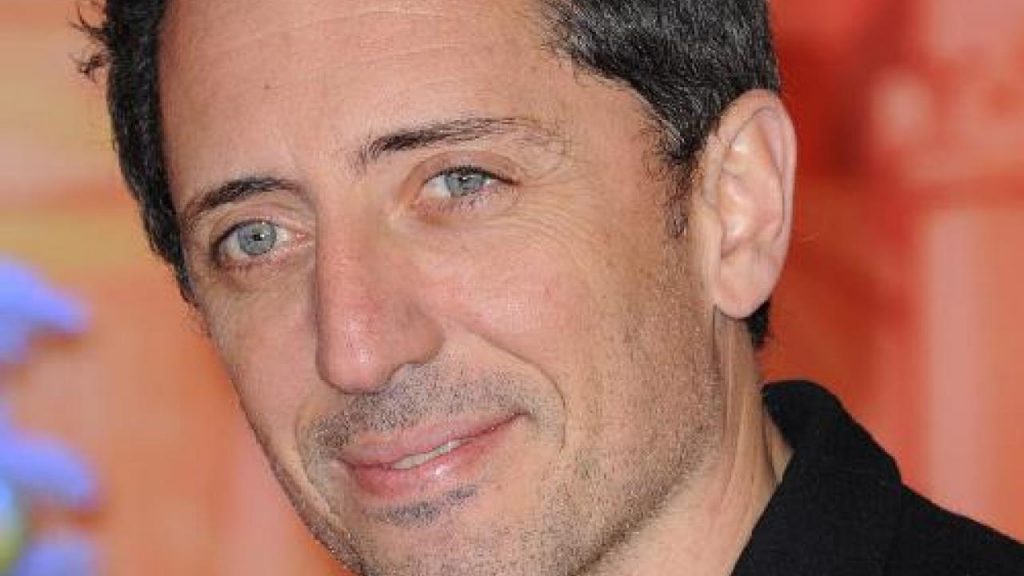 Portrait en gros plan d’un homme aux cheveux courts foncés et aux yeux clairs, affichant un léger sourire, photographié dans un contexte intérieur avec un fond coloré flou.