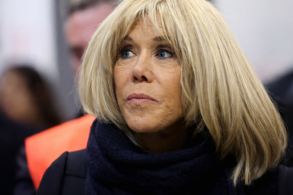 Femme blonde en gros plan, regard concentré, vêtue d’un manteau sombre et d’une écharpe, dans un contexte public flou en arrière-plan