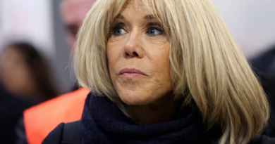 Femme blonde en gros plan, regard concentré, vêtue d’un manteau sombre et d’une écharpe, dans un contexte public flou en arrière-plan