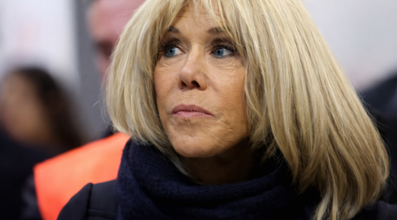 Femme blonde en gros plan, regard concentré, vêtue d’un manteau sombre et d’une écharpe, dans un contexte public flou en arrière-plan