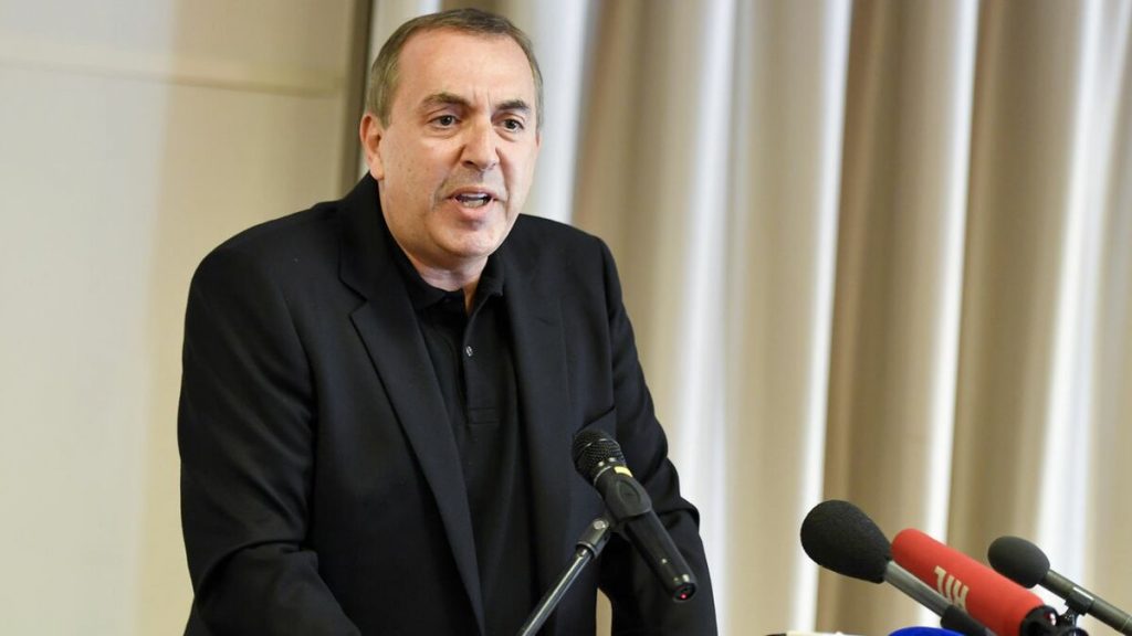 homme s’exprimant devant plusieurs microphones lors d’une prise de parole publique en intérieur