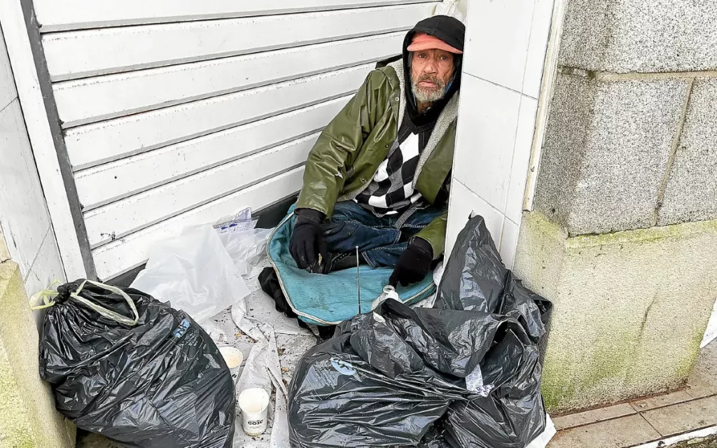 Homme âgé assis au sol sous un porche urbain, entouré de sacs et d’une couverture, exposé à l’humidité et au froid.
