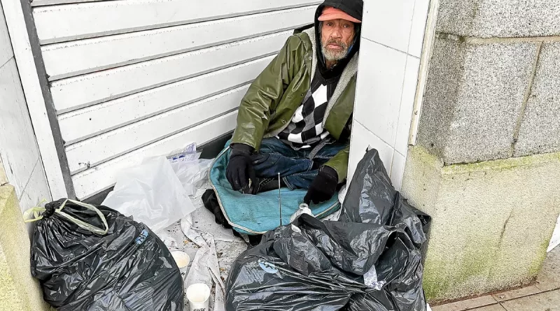 Homme âgé assis au sol sous un porche urbain, entouré de sacs et d’une couverture, exposé à l’humidité et au froid.