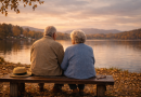 Couple de personnes âgées assises sur un banc en bois face à un lac calme, observant le paysage au coucher du soleil, dans une ambiance automnale et contemplative.