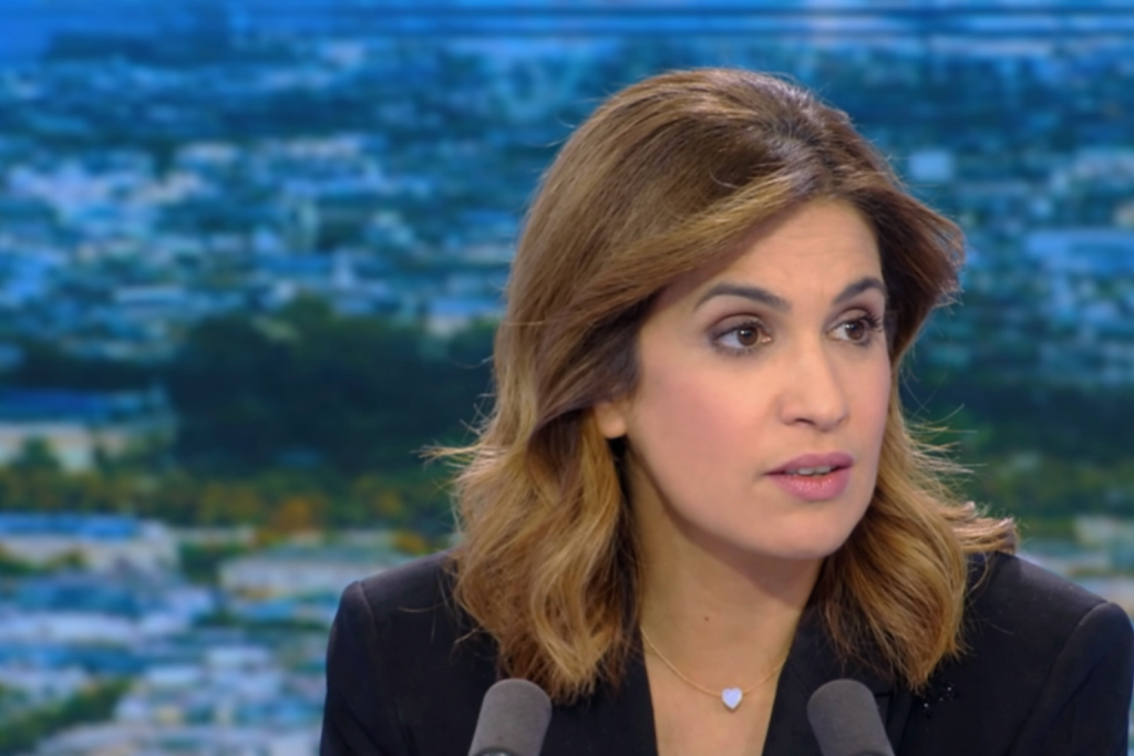 Journaliste de télévision en plateau, regard sérieux, expression grave, microphones visibles au premier plan, décor urbain flouté en arrière-plan.