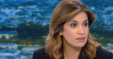 Journaliste de télévision en plateau, regard sérieux, expression grave, microphones visibles au premier plan, décor urbain flouté en arrière-plan.