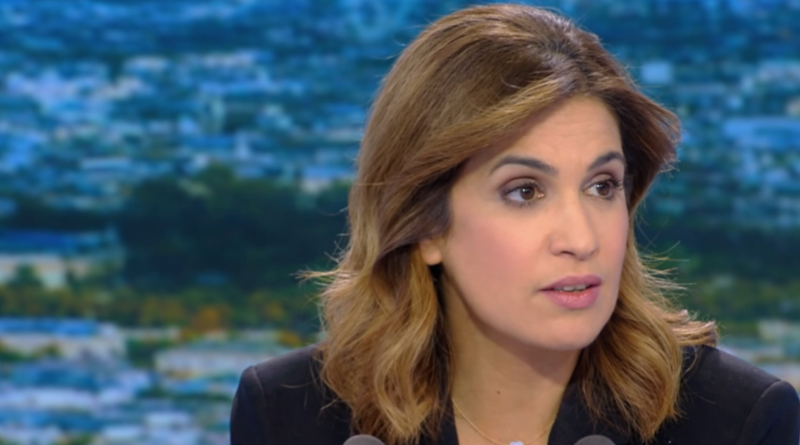 Journaliste de télévision en plateau, regard sérieux, expression grave, microphones visibles au premier plan, décor urbain flouté en arrière-plan.