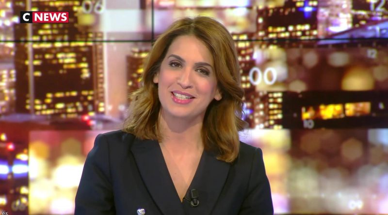 Journaliste femme souriante sur un plateau de télévision, éclairage studio, décor urbain flou en arrière-plan, ambiance chaîne d’information en continu.