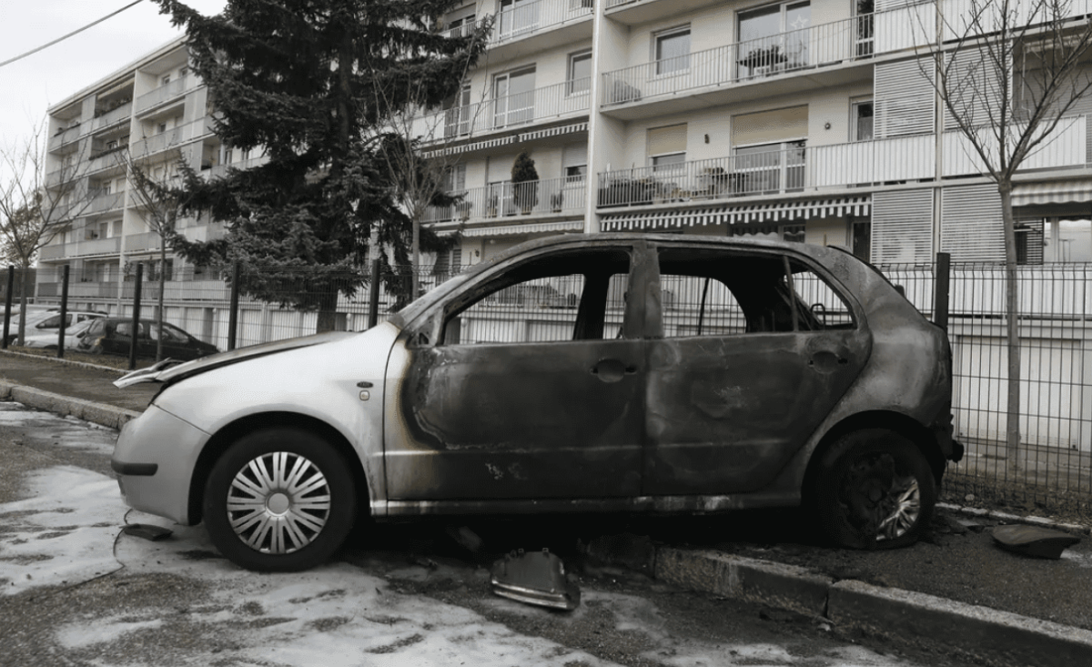 Voiture entièrement calcinée stationnée le long d’un trottoir devant des immeubles résidentiels après un incendie.
