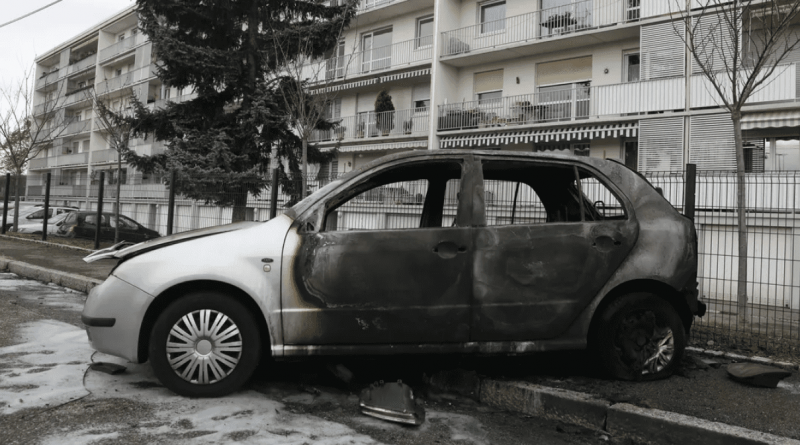 Voiture entièrement calcinée stationnée le long d’un trottoir devant des immeubles résidentiels après un incendie.