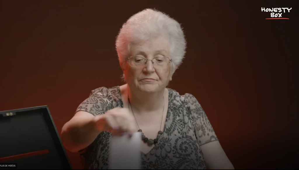 Femme âgée aux cheveux gris courts assise à une table, tendant la main vers un objet, expression concentrée, éclairage tamisé sur fond sombre