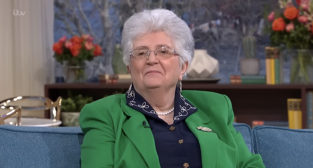 Femme âgée aux cheveux gris courts assise sur un canapé, portant une veste verte et des lunettes, expression calme et posée, décor intérieur lumineux en arrière-plan